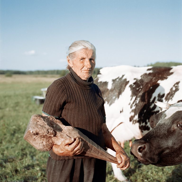 Color photograph portrait by Tadas Kazakevičius, from Soon to be Gone woman holding stick, cow, field
