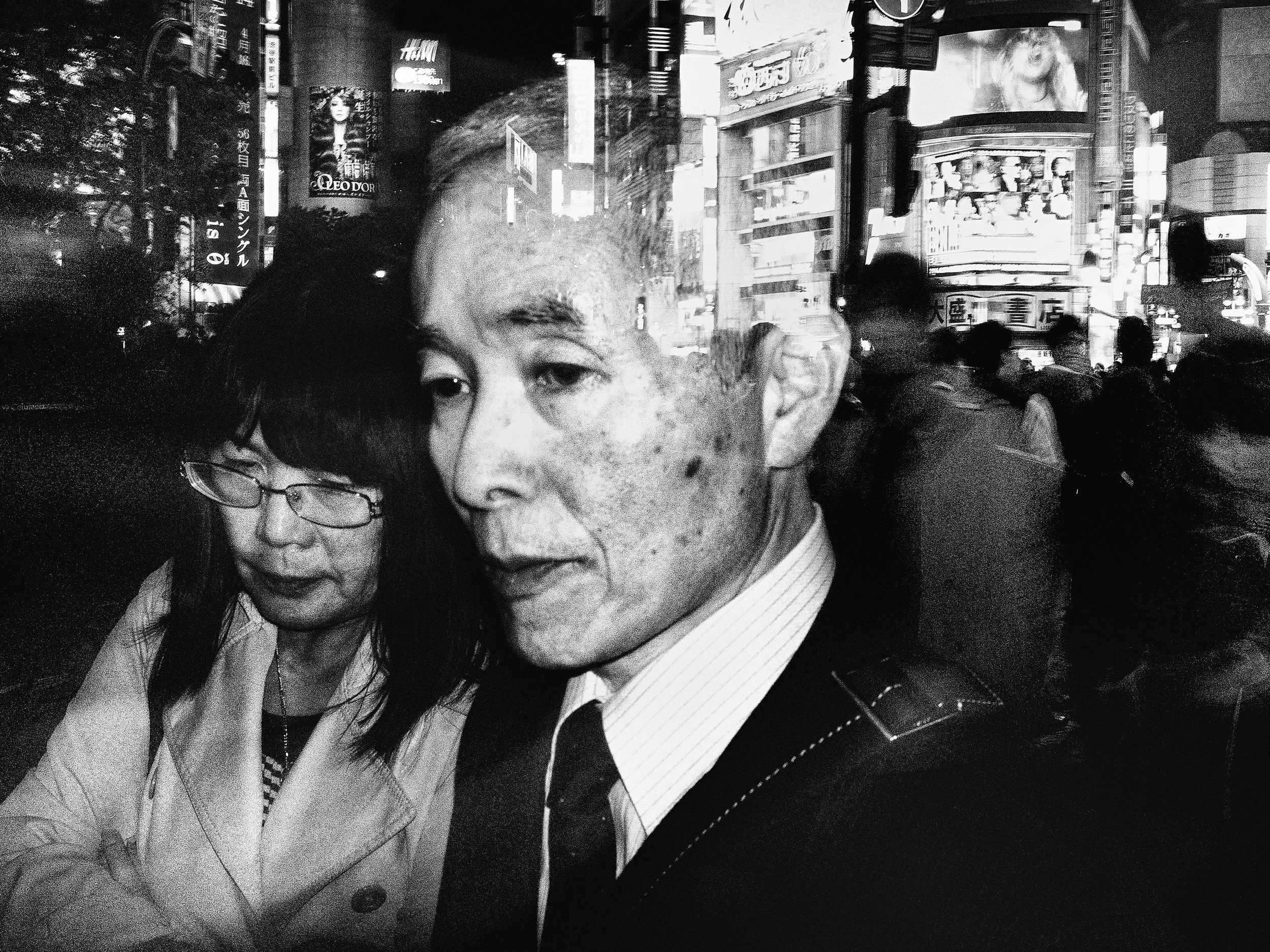 Grainy high-contrast night frame shot from arm's length in a crowd: an elderly man in suit and tie fills two-thirds of the frame, close enough that pores and liver spots read; a woman in glasses presses against his shoulder; behind, a wall of backs dissolving into blown-out neon