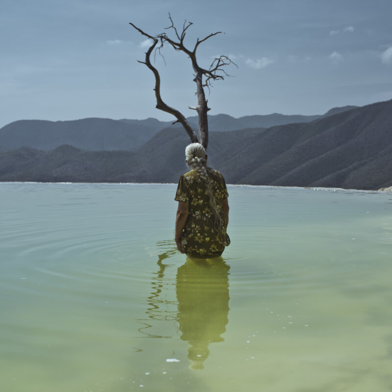 Photo of an elderly woman up to her waist in a lake surrounded by mountains by Cristina de Middel. From the book, Journey to the Center