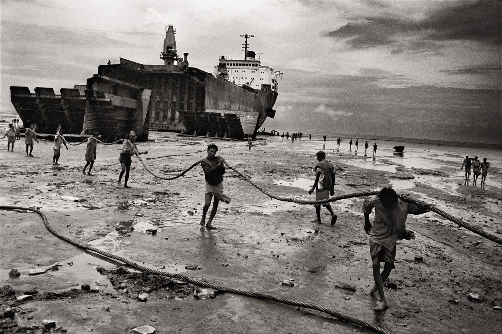 Black & white photography by Ian Berry. Bare-handed men and boys prepare to drag a hawser in the ship-breaking yard. Huge tankers are driven at full throttle and high tide on to the beach where they are broken up mostly by hand with scant regard for health and safety. in Bangladesh