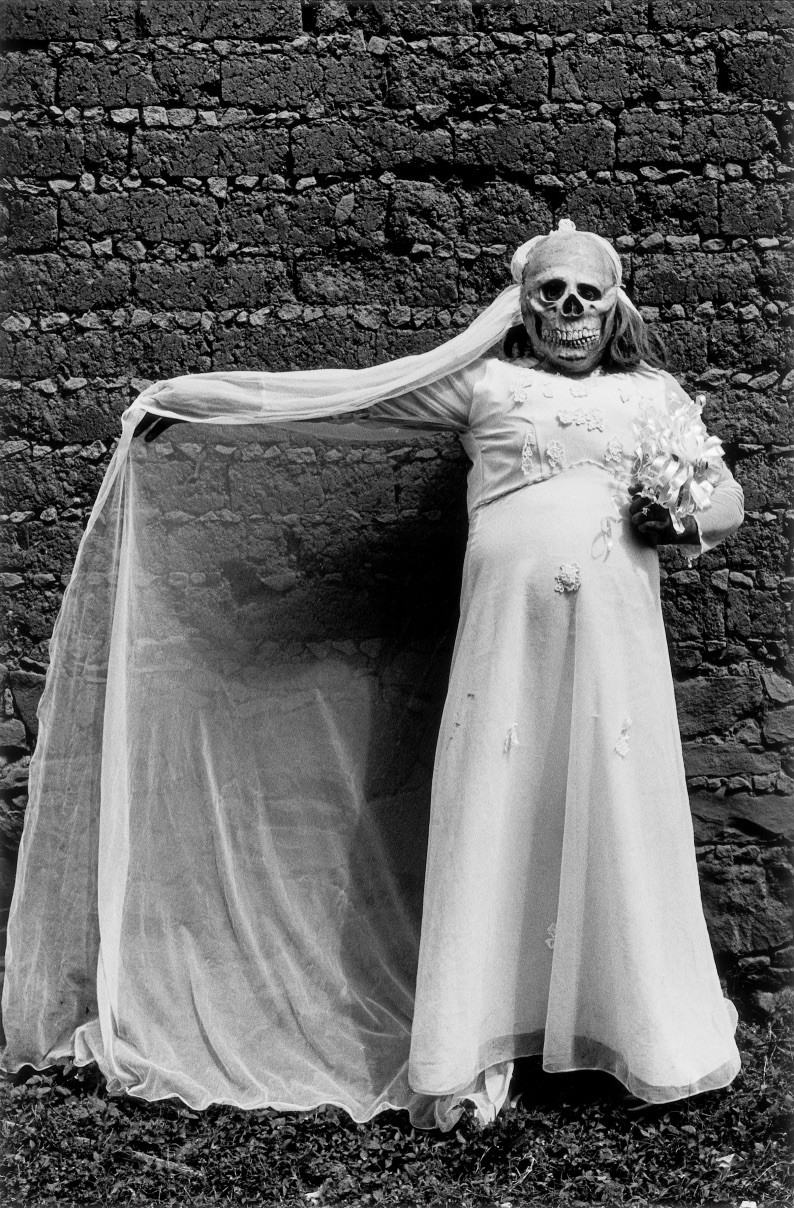 Black & white photography by Graciela Iturbide. Portrait of a woman holding flowers with as skull mask
