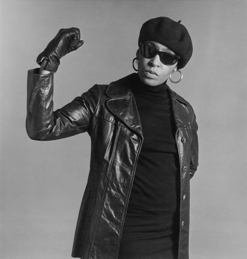 Black and white self portrait by Carrie Mae Weems, dressed in black leather, sunglasses and a beret