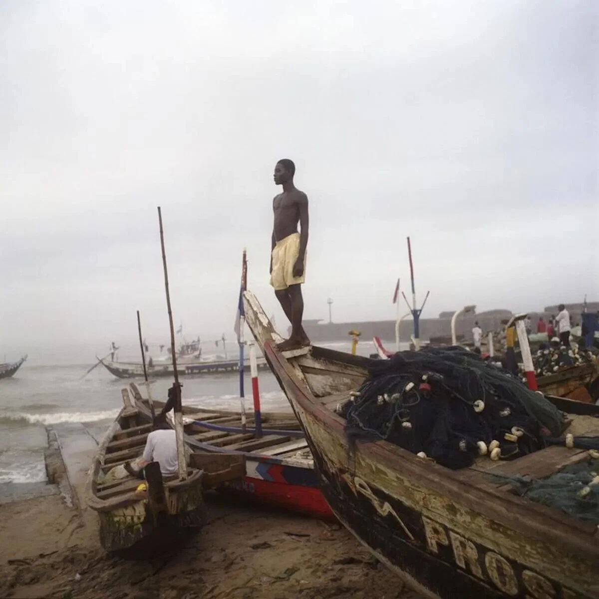 Farbfoto eines Mannes, der auf einem Fischerboot am Ufer in Ghana steht und aufs Meer hinausblickt.