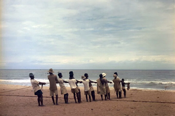 Color photo by Todd Webb of a line of fishermen pulling a line on a beach in Ghana
