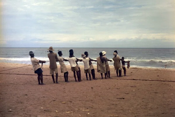 Foto a colori di Todd Webb che ritrae una fila di pescatori intenti a tirare una lenza su una spiaggia del Ghana.