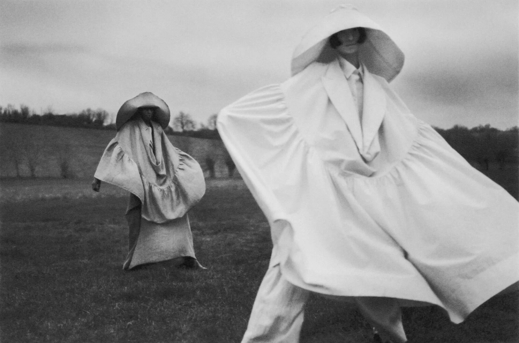 Black & white photography by Amélie Ambroise. Two women in large hats and draped outfits in a field